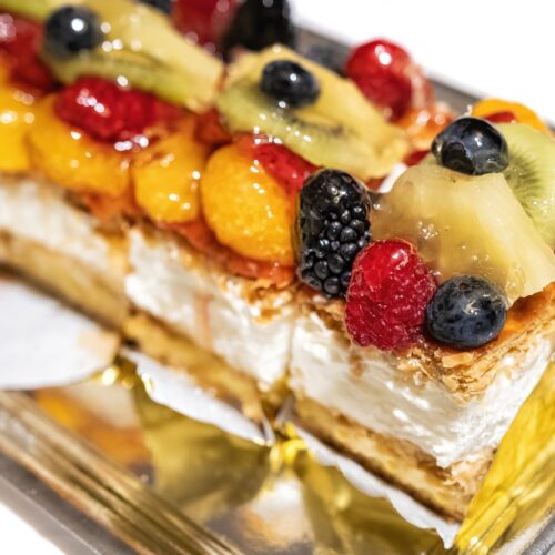 close up of a puff pastry and cream cake with red wild berries, kiwi fruit pieces, pineapple, mandarin slices on a white table, selective focus on one blackberry
